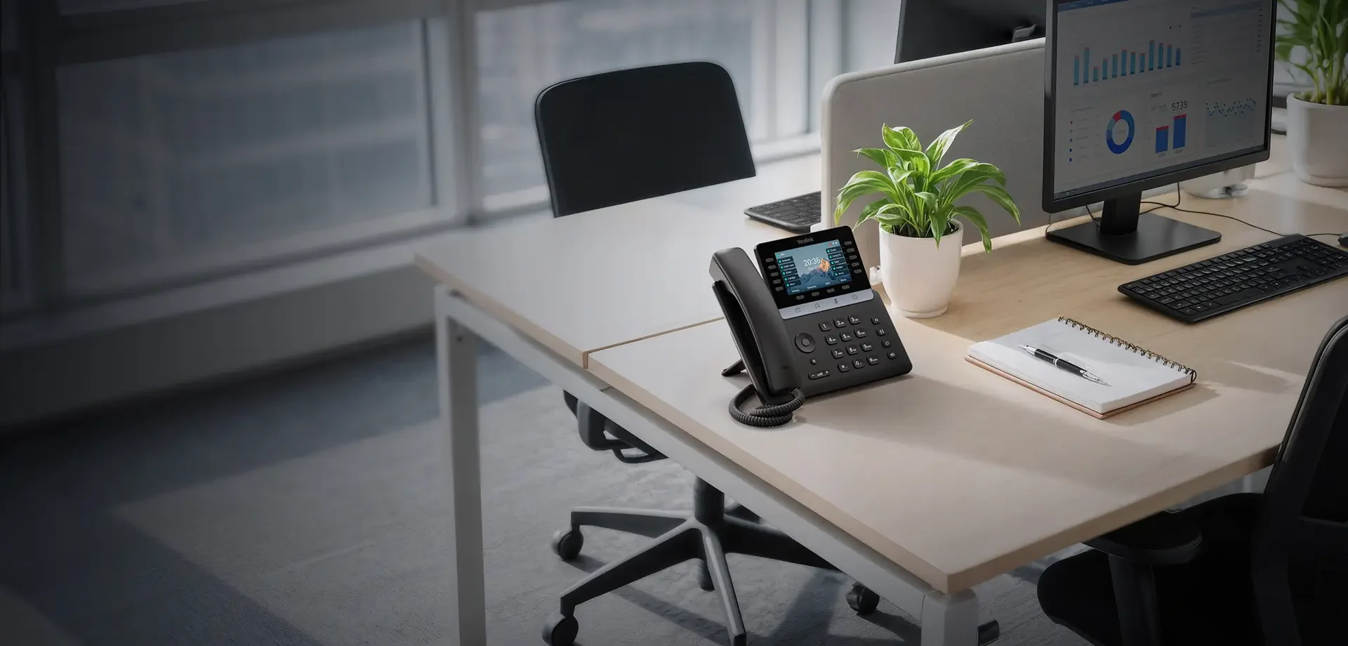A Yealink VoIP desk phone on an office desk beside a monitor showing a live dashboard.